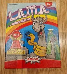 Colorful board game box for L.A.M.A. by Reiner Knizia, featuring a cartoon llama and a rainbow background. The box highlights the game's age recommendation of 8+, its nomination for the Spiel des Jahres award in 2019, and the logo of Amigo Spiele.