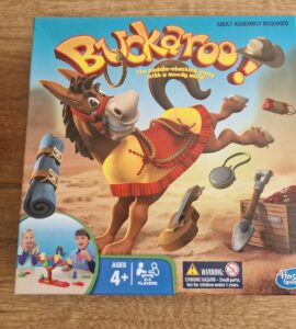 Colorful game box for Buckaroo!, a saddle-stacking game featuring a playful mule, suitable for ages 4 and up, designed for 2-4 players. The packaging displays various game pieces and includes a warning about small parts. Hasbro Gaming branding is visible.