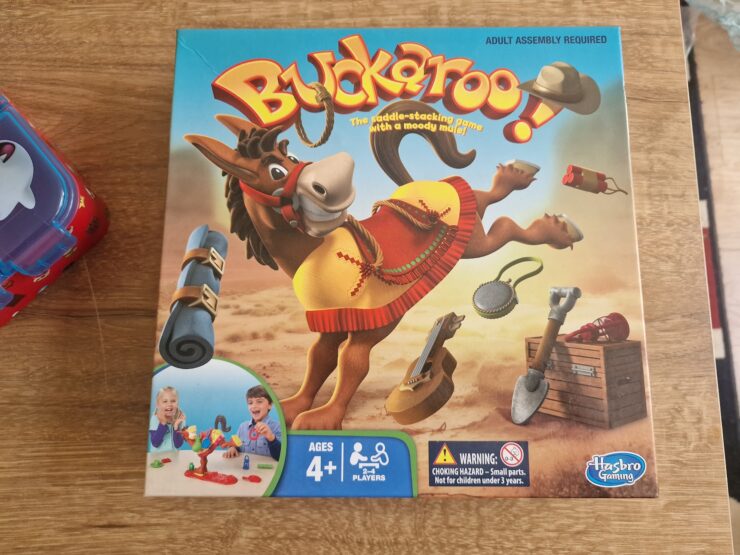 Colorful game box for Buckaroo!, a saddle-stacking game featuring a playful mule, suitable for ages 4 and up, designed for 2-4 players. The packaging displays various game pieces and includes a warning about small parts. Hasbro Gaming branding is visible.