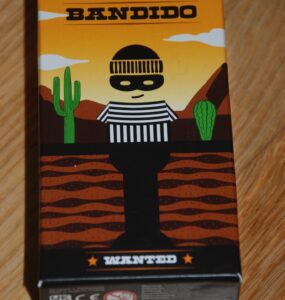 Colorful packaging of the board game "Bandido," featuring a cartoonish bandit character in a striped outfit against a desert backdrop with cacti and mountains. The box is labeled "WANTED" and showcases a playful art style.