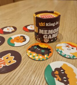 Colorful King Jr. memory game featuring a cylindrical container and various character-themed cards laid out on a wooden table, designed to enhance memory skills and provide family-friendly entertainment.