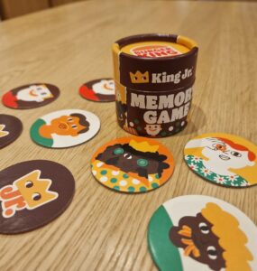 Colorful King Jr. memory game featuring a cylindrical container and various character-themed cards laid out on a wooden table, designed to enhance memory skills and provide family-friendly entertainment.