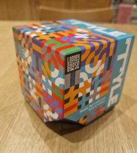 Colorful packaging of the Looney Goose game, featuring a vibrant geometric design and the title "STYLE" prominently displayed on top. The box is positioned on a wooden table, showcasing its unique hexagonal shape and playful artwork.