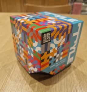 Colorful packaging of the Looney Goose game, featuring a vibrant geometric design and the title "STYLE" prominently displayed on top. The box is positioned on a wooden table, showcasing its unique hexagonal shape and playful artwork.