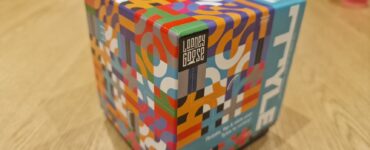 Colorful packaging of the Looney Goose game, featuring a vibrant geometric design and the title "STYLE" prominently displayed on top. The box is positioned on a wooden table, showcasing its unique hexagonal shape and playful artwork.