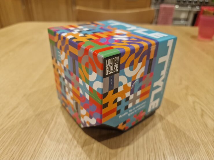 Colorful packaging of the Looney Goose game, featuring a vibrant geometric design and the title "STYLE" prominently displayed on top. The box is positioned on a wooden table, showcasing its unique hexagonal shape and playful artwork.