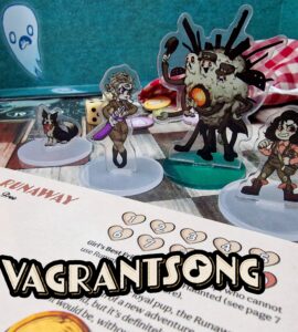 Vagrantsong tabletop game components featuring colorful acrylic character standees, including a dog, a female character with a weapon, and a quirky monster, set against a game board with dice and a character sheet.