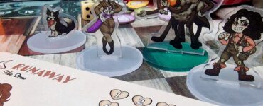 Vagrantsong tabletop game components featuring colorful acrylic character standees, including a dog, a female character with a weapon, and a quirky monster, set against a game board with dice and a character sheet.