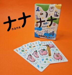 Colorful card game "Nana" by Kaya Miyano and Sai Beppu displayed with its box and several illustrated cards featuring cute animal designs on an orange background.