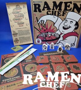 Ramen Chef board game components displayed, including the game box, instruction booklet, ingredient cards, character tokens, and chopsticks on a blue background. The design features vibrant illustrations related to ramen cooking, enhancing the game's culinary theme.