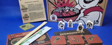 Ramen Chef board game components displayed, including the game box, instruction booklet, ingredient cards, character tokens, and chopsticks on a blue background. The design features vibrant illustrations related to ramen cooking, enhancing the game's culinary theme.