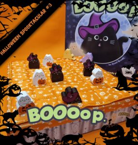 Colorful Halloween-themed board game setup featuring cute cat game pieces, including black and white cats in costumes, on an orange quilted board. The game, titled "Boooop," is designed by Scott Brady, with a festive background of bats and pumpkins. Perfect for family fun during the Halloween season.