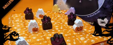Colorful Halloween-themed board game setup featuring cute cat game pieces, including black and white cats in costumes, on an orange quilted board. The game, titled "Boooop," is designed by Scott Brady, with a festive background of bats and pumpkins. Perfect for family fun during the Halloween season.