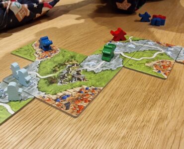 Colorful board game pieces are placed on a wooden table, featuring a landscape of tiles with rivers, cities, and hills. The game involves blue, green, and red player pieces, with several tiles depicting vibrant medieval-style scenery.