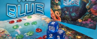 Colorful game setup for "Into the Blue," featuring a vibrant underwater-themed game board, decorative tokens in red and green, and blue dice displaying various numbers. The background highlights the game's title, surrounded by marine life illustrations.