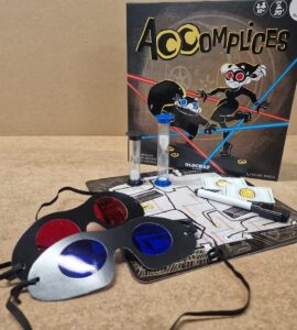 Image of the board game "Accomplices," featuring the game box, playing cards, a timer, colored glasses, and a game board. Ideal for two players aged 10 and above, this game emphasizes strategy and teamwork.