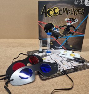 Image of the board game "Accomplices," featuring the game box, playing cards, a timer, colored glasses, and a game board. Ideal for two players aged 10 and above, this game emphasizes strategy and teamwork.