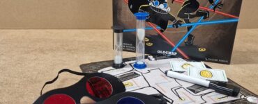 Image of the board game "Accomplices," featuring the game box, playing cards, a timer, colored glasses, and a game board. Ideal for two players aged 10 and above, this game emphasizes strategy and teamwork.