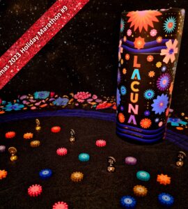 Colorful game pieces scattered on a black fabric surface, featuring the game "Lacuna" prominently displayed. The background showcases vibrant floral designs, and a red banner at the top indicates "Xmas 2023 Holiday Marathon #9." This image highlights a festive and playful atmosphere suitable for game enthusiasts.