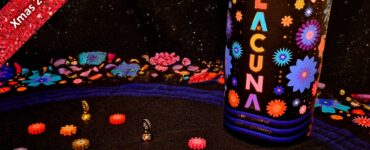 Colorful game pieces scattered on a black fabric surface, featuring the game "Lacuna" prominently displayed. The background showcases vibrant floral designs, and a red banner at the top indicates "Xmas 2023 Holiday Marathon #9." This image highlights a festive and playful atmosphere suitable for game enthusiasts.