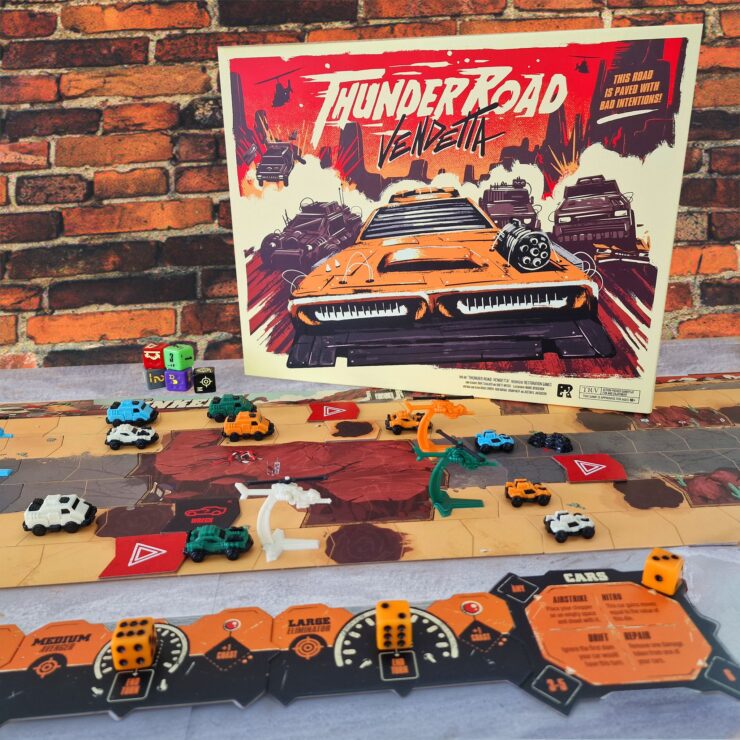 Thunder Road Vendetta board game setup featuring colorful miniature vehicles on a dynamic game board, with the game's vibrant cover art displayed prominently in the background against a brick wall. The scene showcases gameplay elements like dice and player cards, emphasizing the strategic and competitive nature of the game.