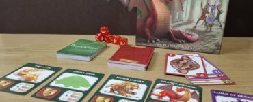 Game board of Dragonwood, a tabletop game featuring colorful cards and dice. The image shows the game box, various character and creature cards, and red dice on a wooden table. Ideal for 2-4 players aged 8 and up, Dragonwood combines strategy and luck in a fantasy setting.