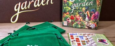 Alice's Garden board game displayed with a colorful box featuring whimsical illustrations, accompanied by several green drawstring bags and game components on a wooden surface.