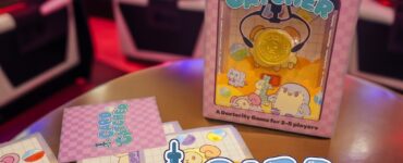 A colorful game box for "Card Catcher," a dexterity game designed for 2-5 players, featuring vibrant illustrations of cute characters and a gold coin. The game is set against the backdrop of arcade machines, highlighting its fun, arcade-inspired theme.