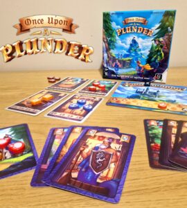 Board game "Once Upon a Plunder" displayed on a wooden table, featuring the game box, character cards, and colorful game tokens. The image showcases gameplay elements, including cards with various illustrations and point values, emphasizing the cooperative and competitive nature of the game.