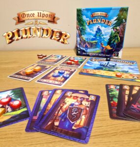 Board game "Once Upon a Plunder" displayed on a wooden table, featuring the game box, character cards, and colorful game tokens. The image showcases gameplay elements, including cards with various illustrations and point values, emphasizing the cooperative and competitive nature of the game.