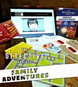 An interactive setup for "The Detective Society: Family Adventures," featuring a laptop displaying a video, game cards, a colorful map, and various clues, all designed for family-friendly mystery-solving activities.