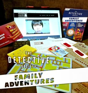 An interactive setup for "The Detective Society: Family Adventures," featuring a laptop displaying a video, game cards, a colorful map, and various clues, all designed for family-friendly mystery-solving activities.