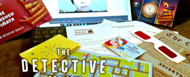 An interactive setup for "The Detective Society: Family Adventures," featuring a laptop displaying a video, game cards, a colorful map, and various clues, all designed for family-friendly mystery-solving activities.