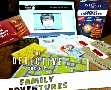 An interactive setup for "The Detective Society: Family Adventures," featuring a laptop displaying a video, game cards, a colorful map, and various clues, all designed for family-friendly mystery-solving activities.