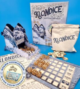 Klondice board game setup featuring the game box, dice, scoring sheet, and a drawstring bag. The game includes a unique mountain-themed design and components for players to stake their claim and earn credits. Ideal for family game nights and strategy enthusiasts.