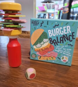 Colorful wooden burger stacking game called "Burger Balance" displayed on a table, featuring a precariously stacked burger made of various toppings and a red bottle, with a colorful die nearby.