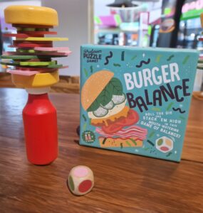 Colorful wooden burger stacking game called "Burger Balance" displayed on a table, featuring a precariously stacked burger made of various toppings and a red bottle, with a colorful die nearby.
