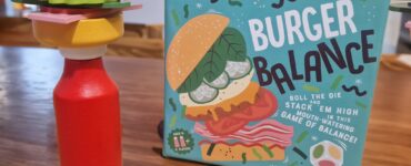Colorful wooden burger stacking game called "Burger Balance" displayed on a table, featuring a precariously stacked burger made of various toppings and a red bottle, with a colorful die nearby.