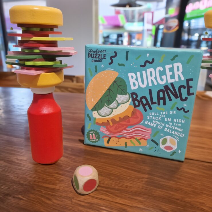 Colorful wooden burger stacking game called "Burger Balance" displayed on a table, featuring a precariously stacked burger made of various toppings and a red bottle, with a colorful die nearby.