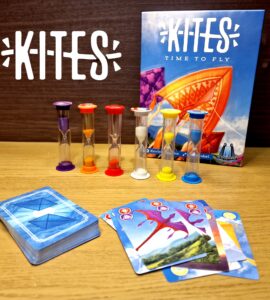 Colorful game components for "Kites: Time to Fly," featuring hourglasses and playing cards on a wooden surface. The game box displays vibrant kite artwork, emphasizing a fun and engaging family game experience.