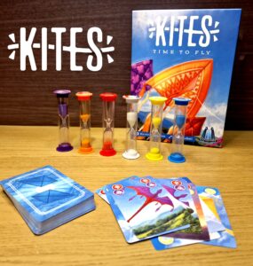 Colorful game components for "Kites: Time to Fly," featuring hourglasses and playing cards on a wooden surface. The game box displays vibrant kite artwork, emphasizing a fun and engaging family game experience.