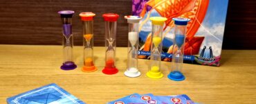 Colorful game components for "Kites: Time to Fly," featuring hourglasses and playing cards on a wooden surface. The game box displays vibrant kite artwork, emphasizing a fun and engaging family game experience.