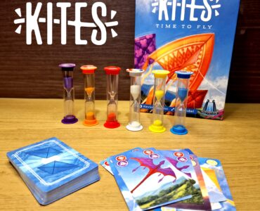 Colorful game components for "Kites: Time to Fly," featuring hourglasses and playing cards on a wooden surface. The game box displays vibrant kite artwork, emphasizing a fun and engaging family game experience.