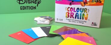 Colour Brain Disney Edition board game displayed on a green background, featuring colorful question cards, answer cards, and a game box with iconic Disney characters. Suitable for 2 to 12 players aged 8 and up, this game challenges players to guess the colors of various Disney-themed items.