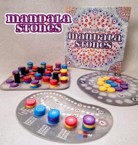 Colorful game components of "Mandala Stones," including various tokens and scoring boards, displayed alongside the game box featuring intricate designs. Ideal for family game nights and strategy lovers.