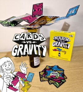 Image of the game "Cards vs Gravity," featuring colorful cards balanced on a bottle, with the game box displayed on a wooden surface. A cartoon character excitedly holds a card, showcasing the fun and challenging nature of this gravity-defying card balancing game.