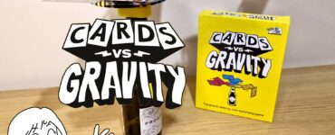 Image of the game "Cards vs Gravity," featuring colorful cards balanced on a bottle, with the game box displayed on a wooden surface. A cartoon character excitedly holds a card, showcasing the fun and challenging nature of this gravity-defying card balancing game.