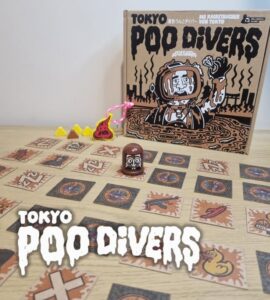 An image depicting the board game "Tokyo Poo Divers," featuring the game box, game pieces, and cards laid out on a wooden surface. The colorful design includes a cartoon diver, playful tokens, and various illustrated cards, showcasing the game's fun and quirky theme.