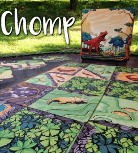 Colorful game board of "Chomp," featuring illustrated dinosaur-themed tiles laid out on a grassy surface, with the game's box displayed in the background.