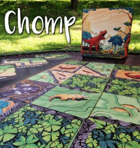 Colorful game board of "Chomp," featuring illustrated dinosaur-themed tiles laid out on a grassy surface, with the game's box displayed in the background.